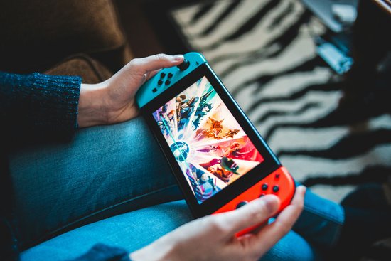 Hände halten eine Nintendo Switch mit blauem und rotem Joy-Con, auf dem Bildschirm ist ein farbenfrohes Spiel mit verschiedenen Charakteren zu sehen.