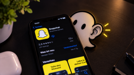 Ein Handy liegt auf einem Tisch. Auf dem Handy ist der App-Store offen und da die Snapchat App als downlaod. Hinter dem Handy guckt der Snapchat Geist raus und lauscht mit seinem Ohr mit.