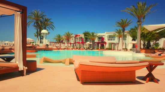 Bild eines in Orange gehaltenen Hotels auf Ibiza. Mehrere Liegen am Pool mit Palmen drumherum.